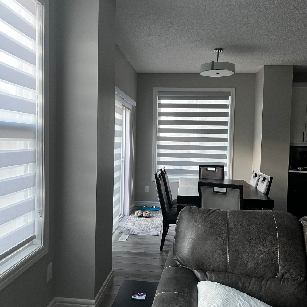 Custom banded shades for an open-concept home. These versatile zebra blinds offer a perfect balance of privacy and light control, complementing a modern interior with gray walls, a dark wood dining set, and leather furniture.