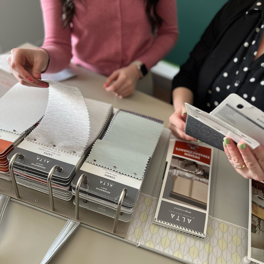 Close-up of a design consultation where a client and consultant are looking through Alta brand fabric samples. One woman lifts a light-textured fabric swatch from a binder, while the other holds a product brochure. Neutral-toned fabric samples and design catalogs are spread across the table.