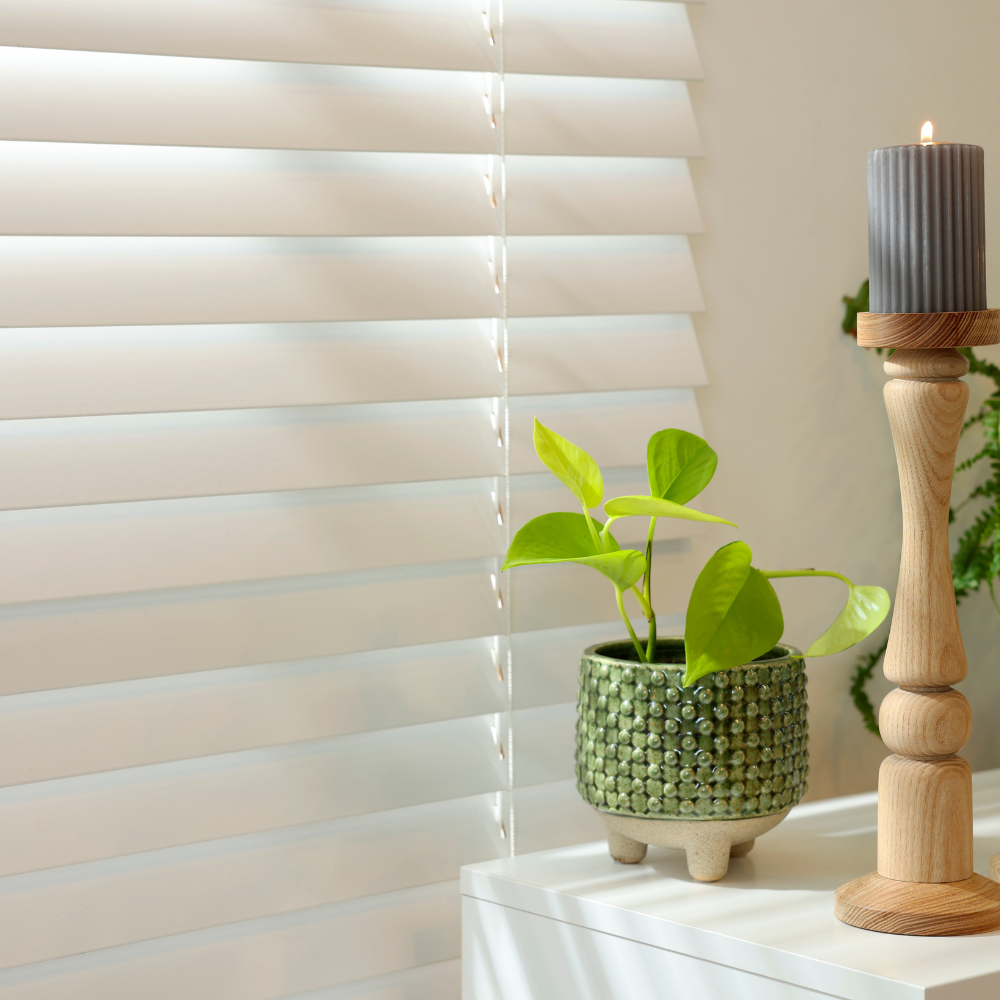 White faux wood blinds filtering soft daylight beside a styled surface with wooden candle holders and green potted plants, creating a clean and cozy home interior scene.