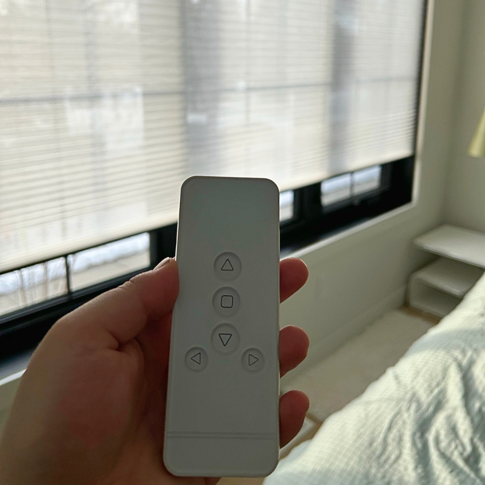 Hand holding a remote control used to operate motorized blinds, with softly filtered light coming through window shades in a modern bedroom setting.